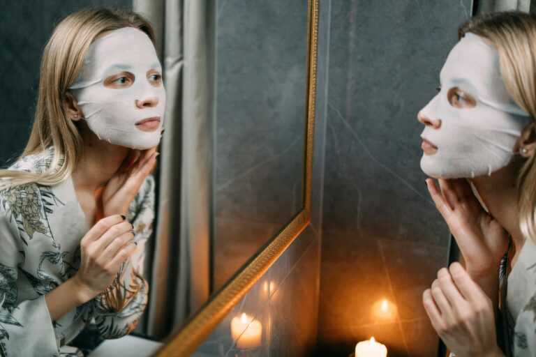 Woman applying a hydrating sheet mask while looking in the mirror, creating a serene and relaxing ambiance.